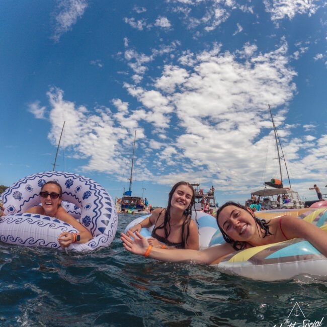 Three women smile and relax on inflatable floaties in the ocean, with boats and people in the background under a blue sky with scattered clouds. A "Yacht Social Club" logo is visible in the bottom right corner.