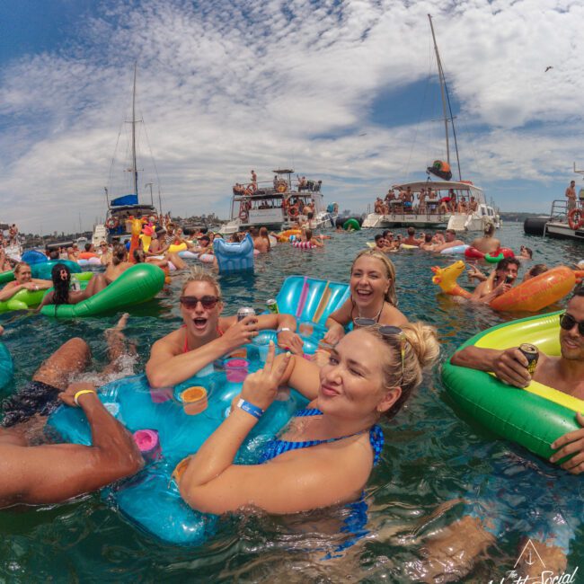 A group of people in swimsuits relax on colorful inflatables in a lively party scene on the water, with boats and more people in the background under a partly cloudy sky.