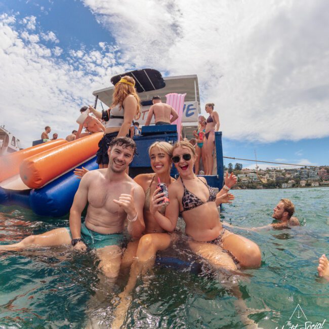 Three people in swimsuits smile and pose on the edge of a boat in the water, with more people behind them enjoying a sunny day, slides, and floating. The scene is lively and festive under a partly cloudy sky.