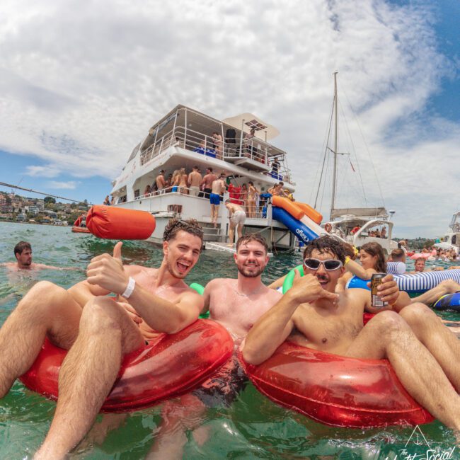 Three young men in swim trunks sit on red inflatables, smiling and posing in front of a crowded party boat on the water. Other people on floats and boats are visible under a sunny sky.