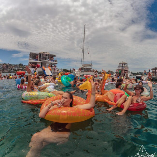 A group of people relax on colorful inflatable pool floats in the water, enjoying drinks and socializing. Boats and yachts are anchored in the background under a partly cloudy sky. "Yacht Social Club" logo is visible.