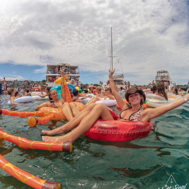 A woman in a cowboy hat smiles and poses on a red inflatable float in the ocean, surrounded by other people on colorful inflatables. Yachts and a party platform are visible in the background under a partly cloudy sky.