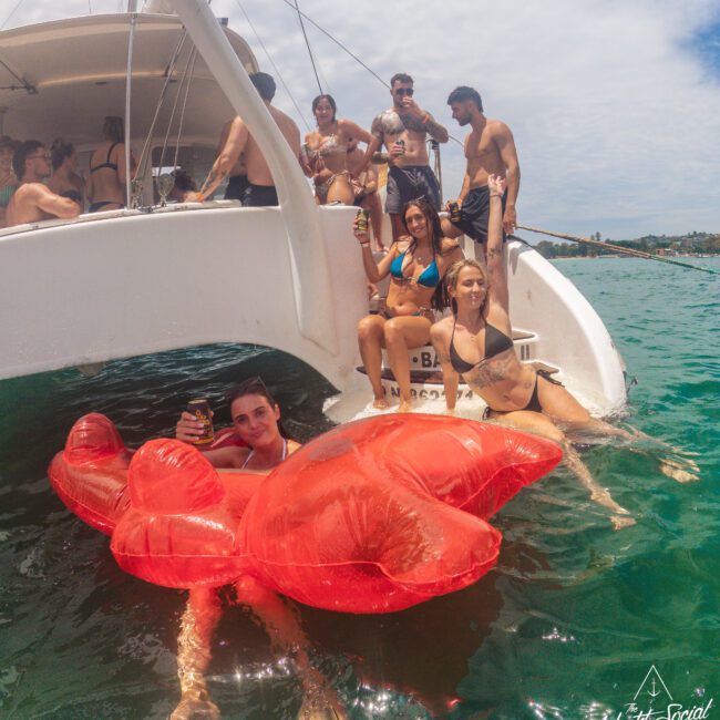 A group of young adults in swimsuits relax on a boat and swim in the sea, with one person floating on a large red inflatable lobster. The sky is partly cloudy and the mood is festive and cheerful.