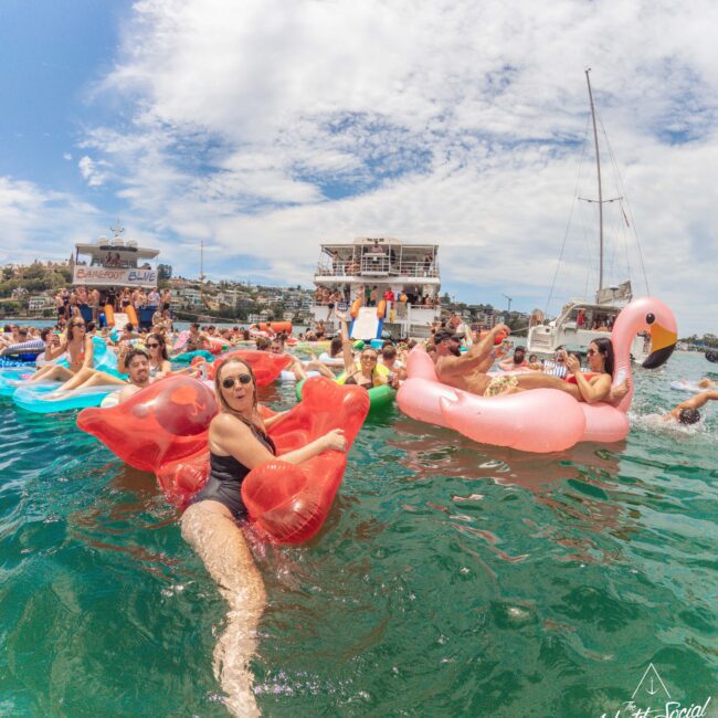 People relax on colorful inflatable floats, including a large pink flamingo, in the water near boats during a sunny outdoor party. The atmosphere is lively, with swimmers and partygoers enjoying the event.