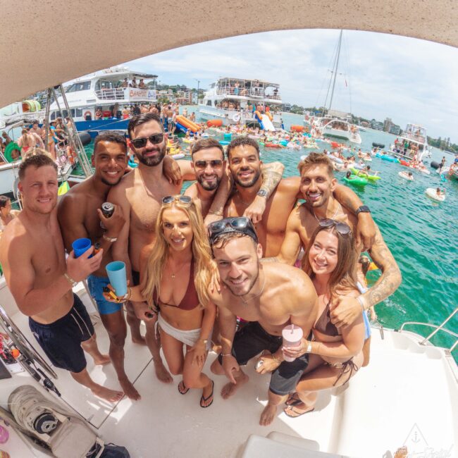 A group of smiling friends in swimsuits pose on a boat, holding drinks, with a busy lake in the background filled with boats, people, and colorful floaties on a sunny day.