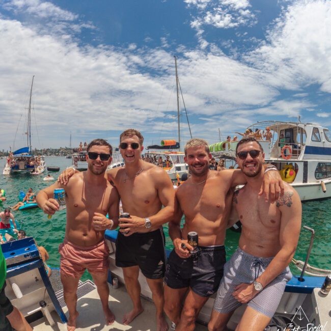 Four smiling men in swim trunks stand arm in arm on a boat, holding drinks. Boats and people are visible on the water under a partly cloudy sky, creating a lively party atmosphere.