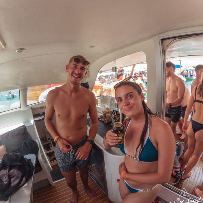 Two people in swimwear smile at the camera inside a boat, each holding a drink. Other people are visible outside through the boat’s windows, enjoying a sunny social gathering on the water.