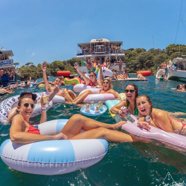A group of smiling people relax on colorful pool floats in the water, holding drinks and posing for the camera. Boats and more people are in the background, with trees lining the shore under a sunny sky.