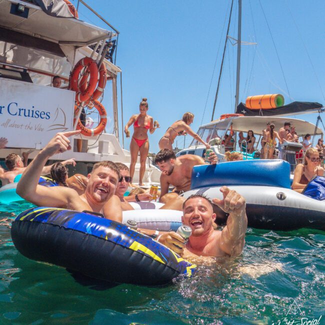 A group of people enjoy a lively pool party near boats on a sunny day. Two men in front smile and gesture while floating on inflatable rings in the water; others relax on floats and dance on the boat decks in the background.