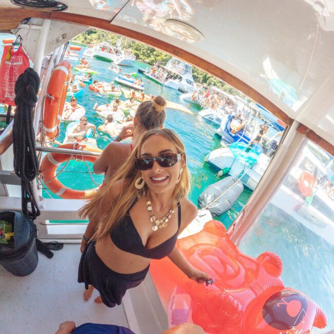 A smiling woman in sunglasses and a black bikini stands on a boat’s deck, holding a red float. Other people, boats, and colorful floats fill the water behind her, creating a lively summer party scene.