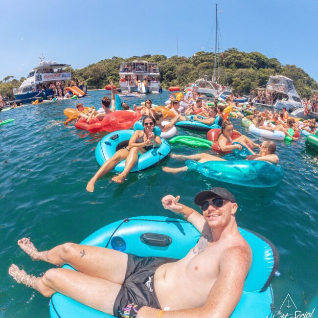A group of people relax on colorful inflatable tubes in the water near boats, enjoying a sunny day. The scene is lively, with many smiling faces and blue skies overhead.