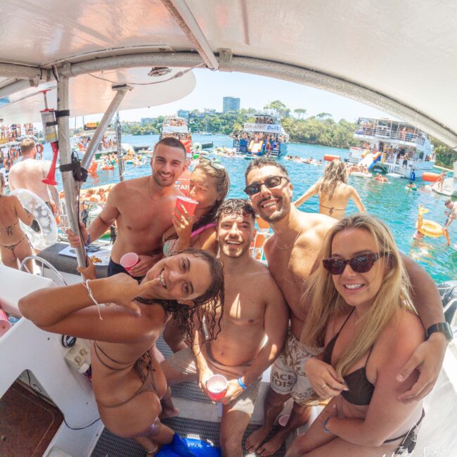 A group of six smiling people in swimsuits pose for a photo on a boat, holding drinks. Behind them, many other boats and people enjoying a sunny day on the water are visible. The atmosphere is festive and lively.