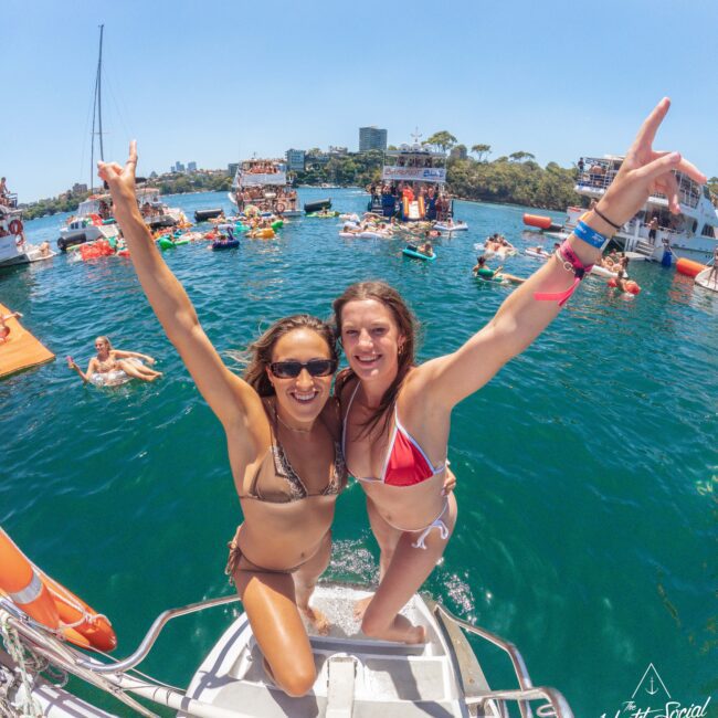 Two women in bikinis smile and pose with arms raised on the back of a boat, with a lively party on the water and other boats in the background under a sunny sky. Logo "Yacht Social Club" appears in the corner.