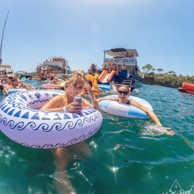 Two women smiling and relaxing on inflatable rings in clear blue water near boats, with more people swimming and enjoying a sunny day. In the background is a party boat with slides and people onboard.
