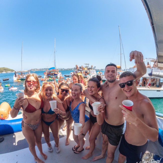 A group of smiling young adults in swimwear hold drinks and pose on a boat with other boats and people on the water in the sunny background, enjoying a lively summer party.