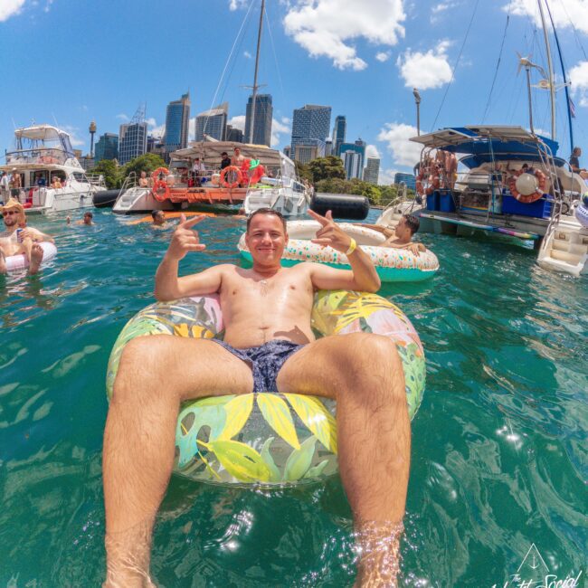 A man lounges on a colorful inflatable ring in the water, making hand signs, with boats and city skyscrapers in the background under a sunny, blue sky. Other people are swimming and relaxing nearby.