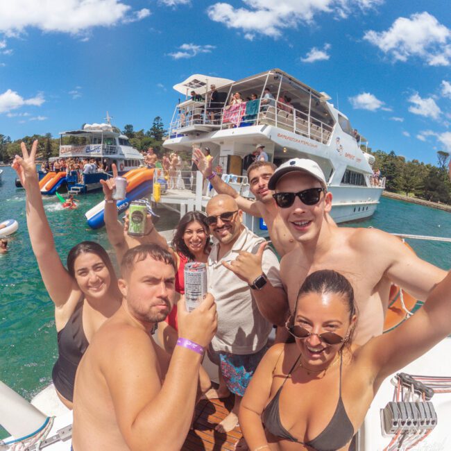 A group of smiling people in swimsuits pose for a selfie on a boat with drinks, surrounded by clear blue water and other party boats in the background under a sunny, partly cloudy sky.