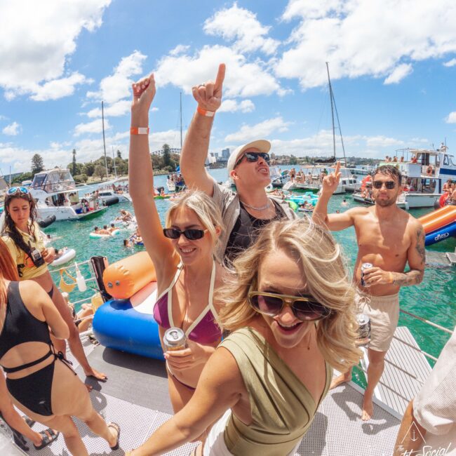 A group of people dance and smile on a docked boat at a lively outdoor party, surrounded by others on nearby boats in clear turquoise water under a sunny, blue sky.