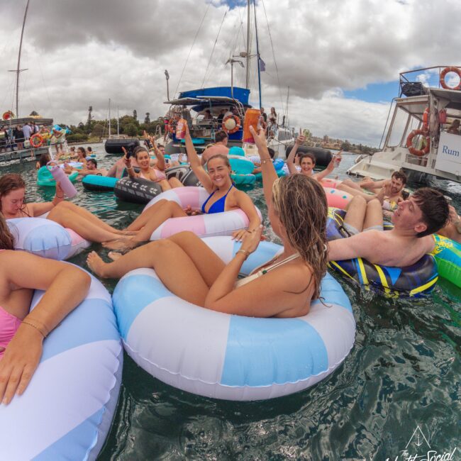 A group of people float on colorful inflatable rings in the water near boats, smiling and raising their arms as they enjoy a lively social gathering under a cloudy sky.