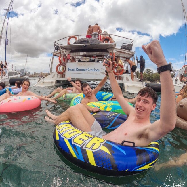 Young men in inflatable tubes smile and cheer while floating in the water near a boat labeled "Rum Runner Cruises." Other people relax in the water and on the boat, enjoying a lively social gathering.