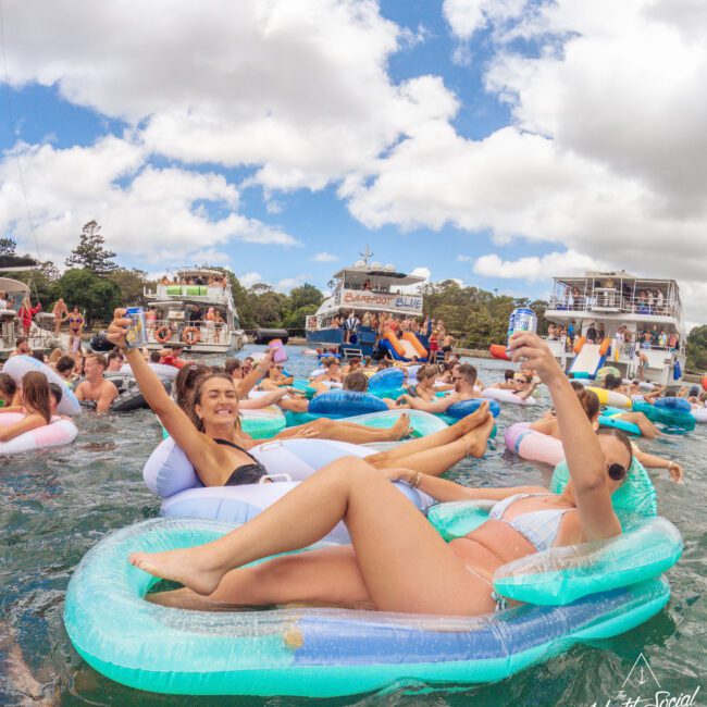 A group of people relax and have fun on colorful inflatable pool floats in the water near several boats, smiling and raising their drinks under partly cloudy skies. The atmosphere is lively and festive.