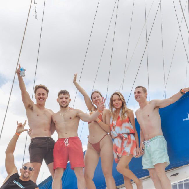 Five smiling young adults in swimwear stand on a boat with blue sails, posing and waving at the camera under a cloudy sky. Two other people are partially visible at the bottom left of the photo.