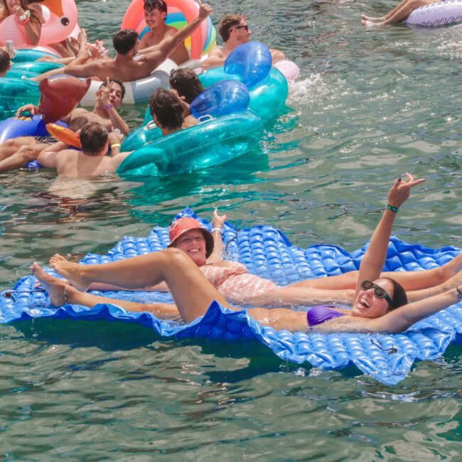 Two women relax and smile on a blue raft in a lake, surrounded by other people on colorful pool floats. The water is calm and everyone appears to be enjoying a sunny day.