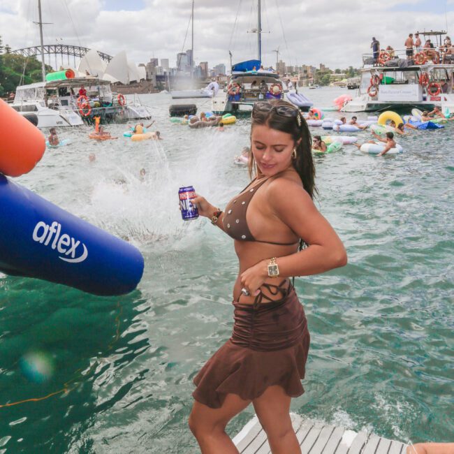 A woman in a brown bikini and skirt holds a drink can on a boat dock. Behind her, people swim and float on pool toys near yachts, with a cityscape and cloudy sky in the background.