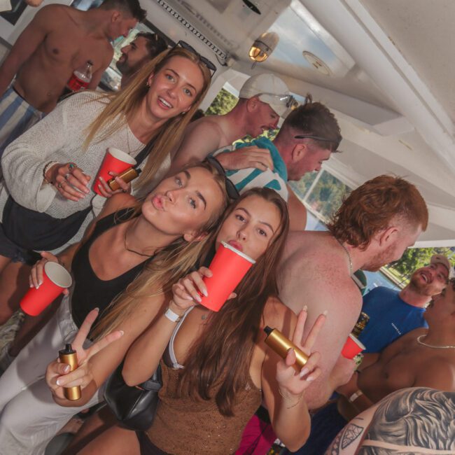 A group of young people at a lively boat party, some holding red cups and smiling or making peace signs, with others mingling in swimwear in the background.