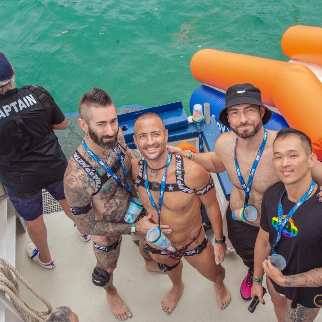 Four smiling men in swimwear and event lanyards stand on a boat near a pool float, posing for a photo. A person in a "Captain" shirt faces away at the edge of the boat above turquoise water.