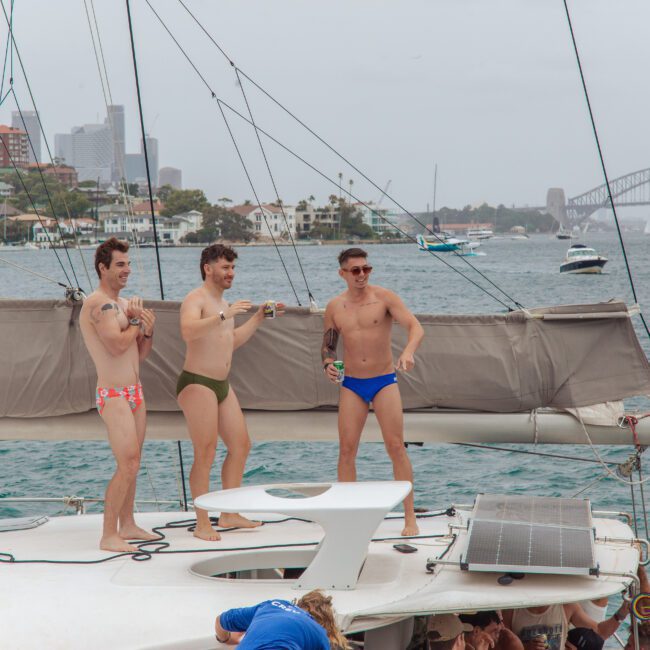 Three men in colorful swim briefs stand on a yacht, laughing and holding drinks, with a city skyline, boats, and a bridge visible in the background on a cloudy day.