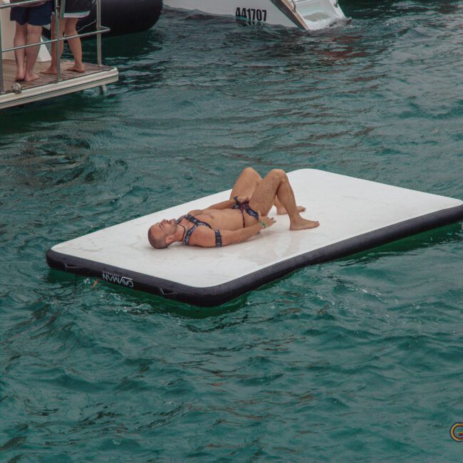 A person in a swimsuit is lying on their back on a floating white mat in green water, with several people standing on a nearby boat in the background. The scene looks relaxed and summery.
