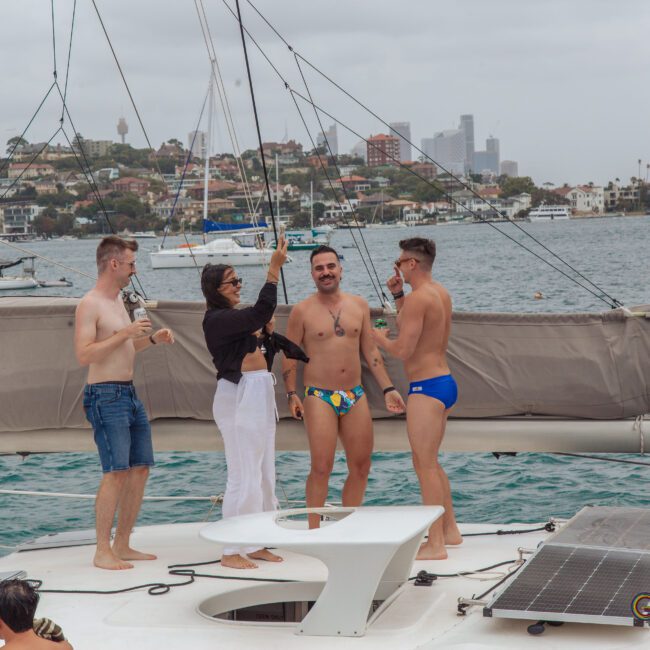 Four people stand on a yacht deck, smiling and toasting drinks, with a city skyline and waterfront houses in the background on an overcast day. Two men wear swim briefs, one man wears shorts, and a woman is dressed in black and white.