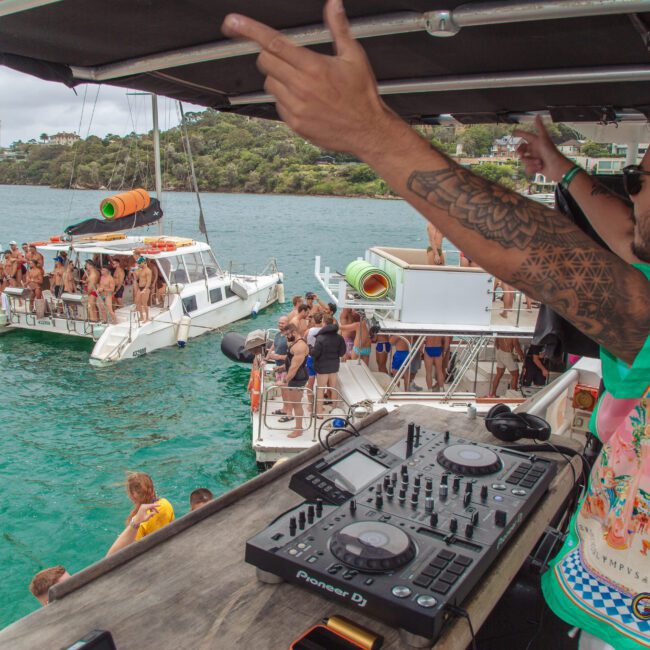 A DJ in a colorful shirt plays music on a boat while people dance and socialize on nearby boats in a scenic bay. The atmosphere is lively and festive with clear water and greenery in the background.