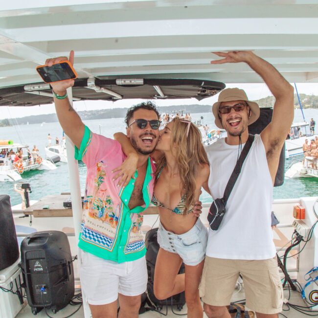Three friends pose happily on a boat; one woman in a bikini kisses a man wearing sunglasses and a colorful shirt, while another man in a hat and sunglasses smiles and raises his arm. Other boats and people are visible in the background.