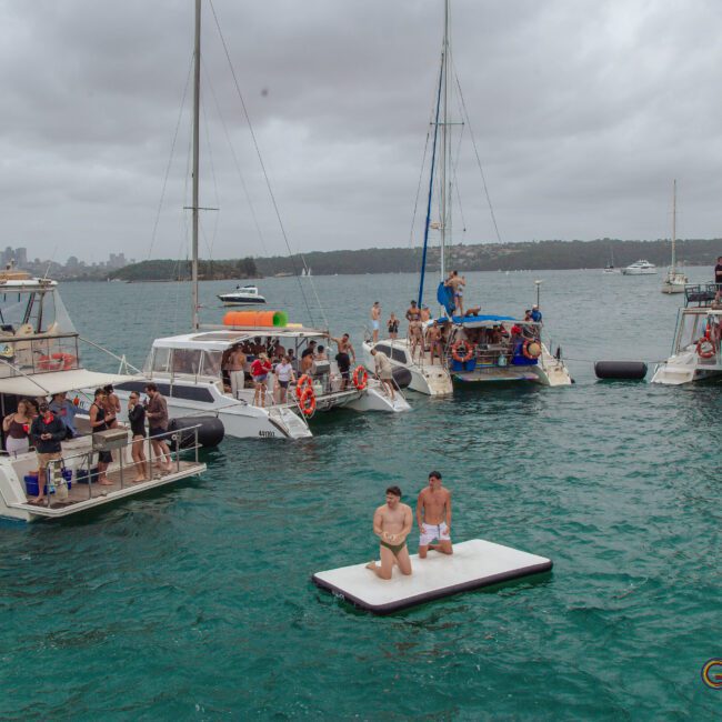 Several boats with groups of people are anchored close together on a cloudy day. Two men are kneeling on a floating platform in the water between the boats. The atmosphere appears festive and lively.