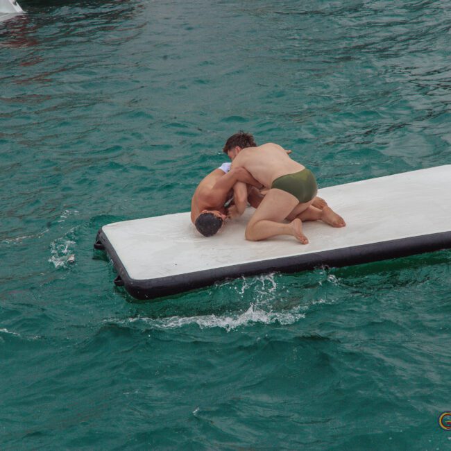 Two people in swimwear wrestle on a floating white mat in the water, surrounded by turquoise waves, near boats.