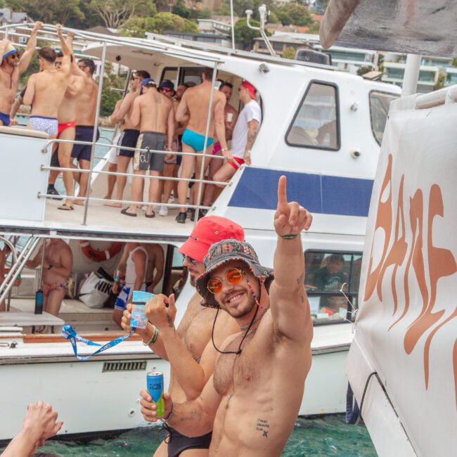 A group of men in swimwear are dancing and celebrating on boats in the water; one man in blue briefs and a bucket hat smiles and points upward, holding a drink, while others party in the background.