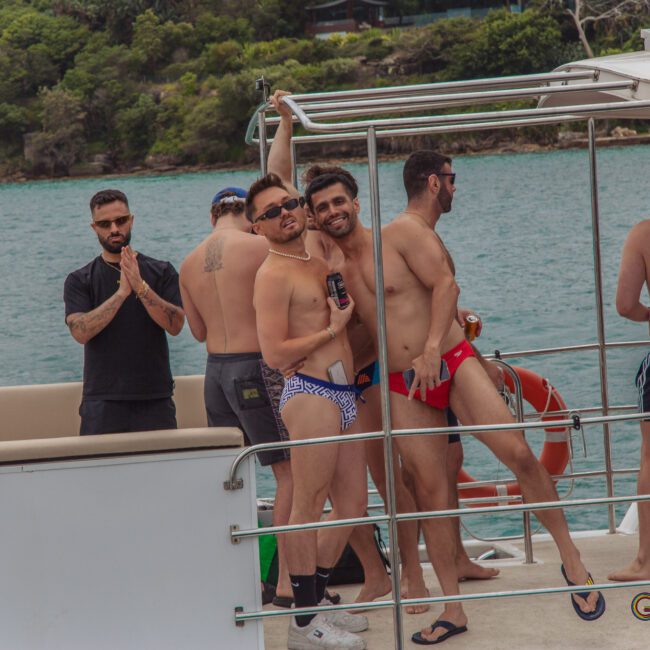 A group of men in swimwear are gathered on a boat, some smiling and posing playfully for the camera, with water and trees visible in the background.