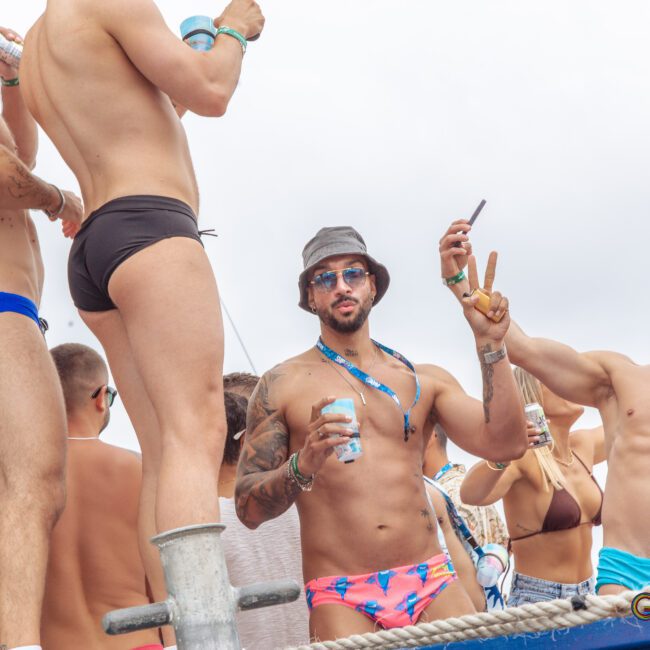 A group of men in swimwear are standing on a boat, socializing, taking selfies, and holding drinks during a lively outdoor event. The atmosphere is festive and relaxed under a bright sky.