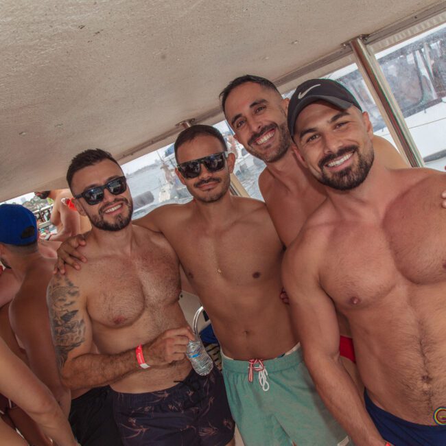 Four smiling, shirtless men wearing swim trunks and sunglasses pose together on a boat with other people in the background, enjoying a sunny day.