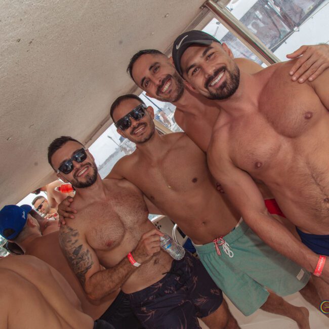 Four smiling men wearing swim trunks and sunglasses pose closely together on a boat, surrounded by other people. They appear to be at a sunny, lively pool or boat party.