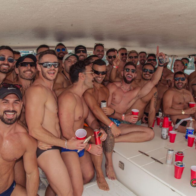 A large group of men in swimwear smiles and poses together on a boat, holding drinks and red cups. The atmosphere is lively and festive, with sunlight filtering in and drinks on a table.
