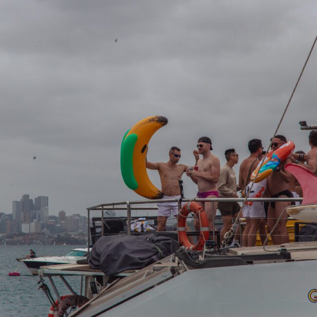 A group of men in swimwear stand on a yacht holding colorful inflatable pool toys, including a banana and a donut, against a cloudy sky with a city skyline in the background.