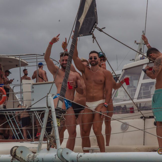 A group of men in swimwear smile, pose, and hold drinks on a crowded boat during a party. Other people are visible in the background, with overcast skies above.