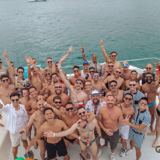 A large group of men in swimwear and sunglasses smile and pose together on a boat, with the ocean and other boats visible in the background. The atmosphere is lively and joyful. GAYM and Lucca Visual logos appear in the corner.