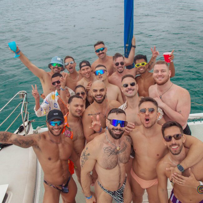 A group of smiling men in swimwear pose together on a boat, holding drinks and making peace signs, with the ocean in the background. The mood is festive and relaxed.