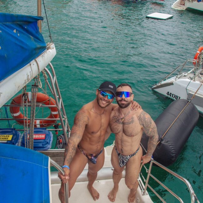 Two men in swimwear and sunglasses stand on the steps of a boat, smiling and posing together. The boat is on turquoise water, with other boats and people in swimwear visible in the background.