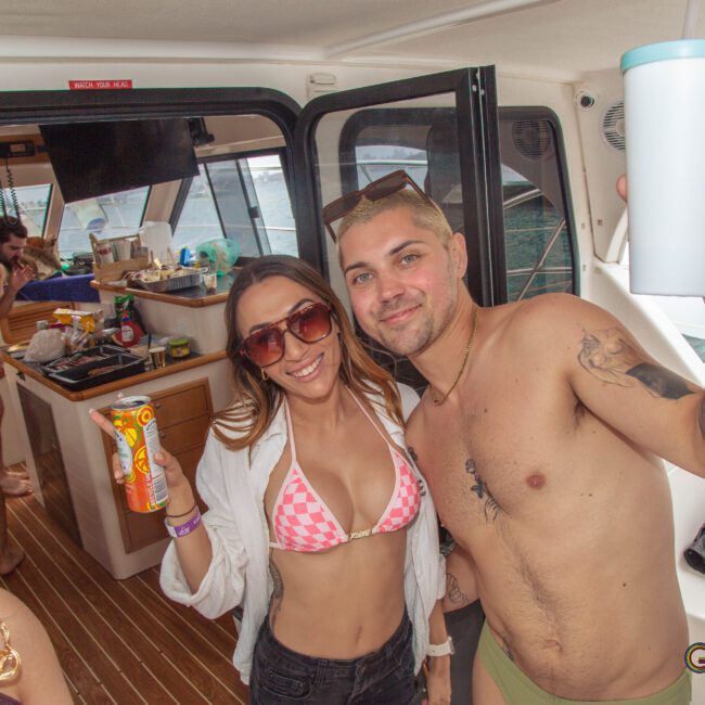 Two people in swimsuits smile and pose with drinks on a boat, surrounded by others enjoying a party. The background shows a kitchen area and open doors to the outside.