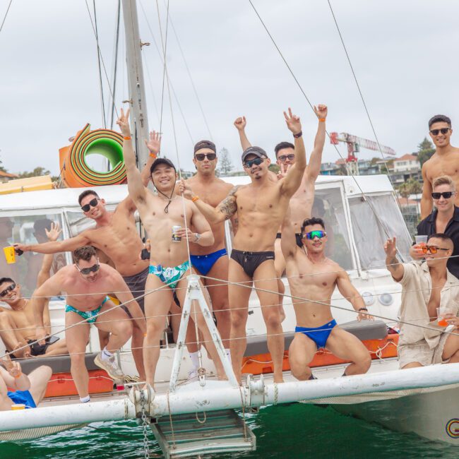 A group of men in swimwear are smiling, raising their arms, and posing on the deck of a boat during a lively outdoor gathering on the water. Some are wearing sunglasses, and the mood appears festive and joyful.